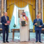 Turkish President Recep Tayyip Erdoğan, Saudi Crown Prince Mohammed bin Salman, and Pakistani Prime Minister Shehbaz Sharif stand side by side holding signed folders after the Mecca Joint Defense Agreement ceremony in a formal hall with yellow curtains and city views, August 7, 2026.