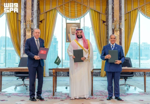 Bangladesh Moves Toward the Saudi-Pakistan Military Alliance: New Delhi on High Alert Turkish President Recep Tayyip Erdoğan, Saudi Crown Prince Mohammed bin Salman, and Pakistani Prime Minister Shehbaz Sharif stand side by side holding signed folders after the Mecca Joint Defense Agreement ceremony in a formal hall with yellow curtains and city views, August 7, 2026.