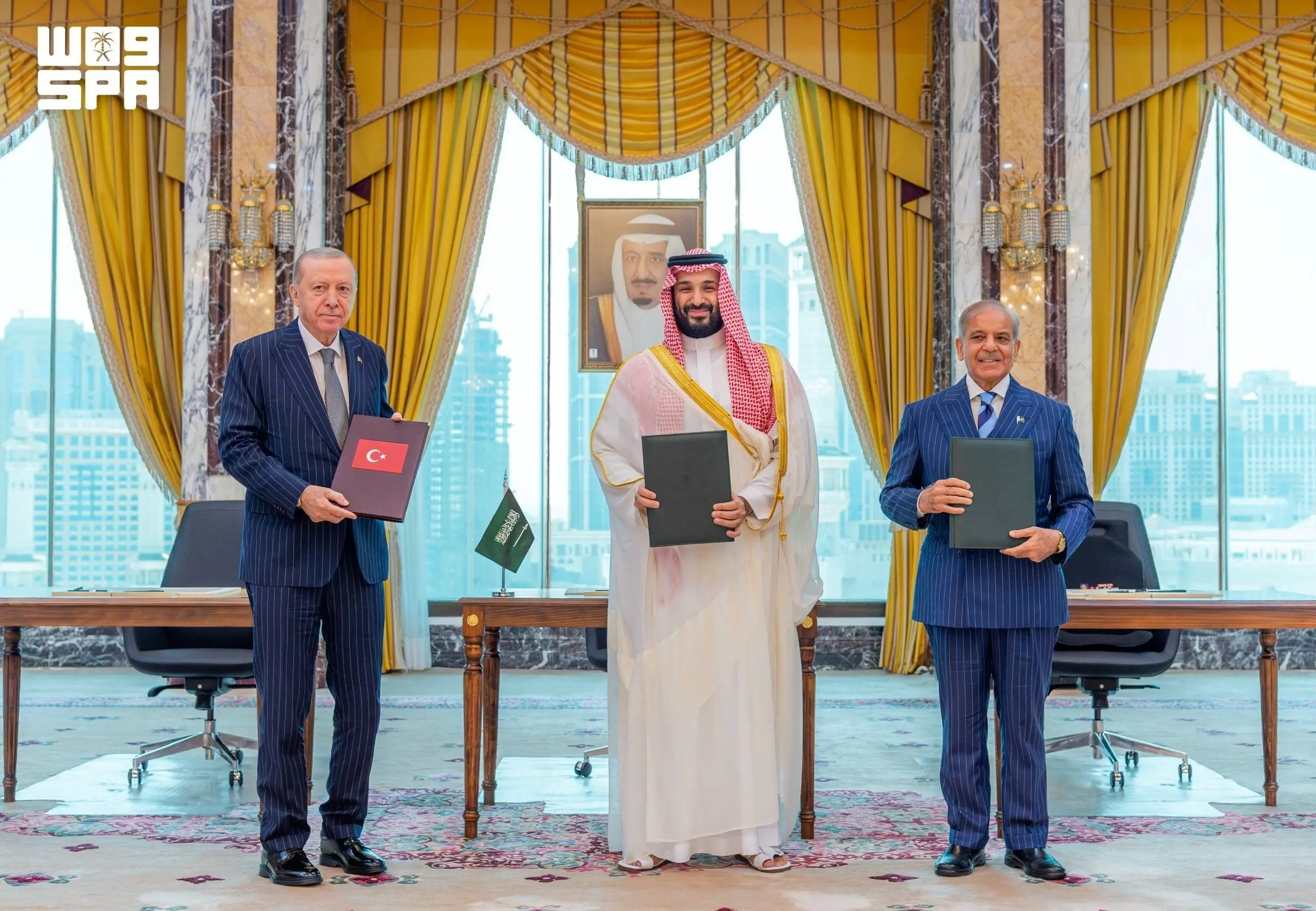 Turkish President Recep Tayyip Erdoğan, Saudi Crown Prince Mohammed bin Salman, and Pakistani Prime Minister Shehbaz Sharif stand side by side holding signed folders after the Mecca Joint Defense Agreement ceremony in a formal hall with yellow curtains and city views, August 7, 2026.