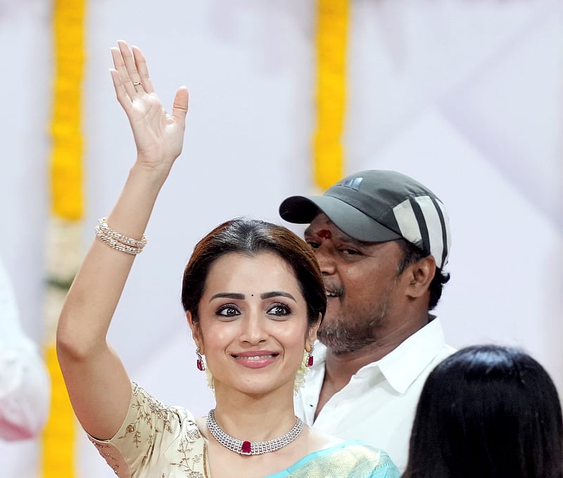 A close-up of actress Trisha Krishnan smiling and waving to supporters at the Jawaharlal Nehru Stadium in Chennai. She is wearing an ice-blue Kanjeevaram silk saree with a cream-embroidered blouse, a diamond and ruby choker, and jasmine flowers in her hair.