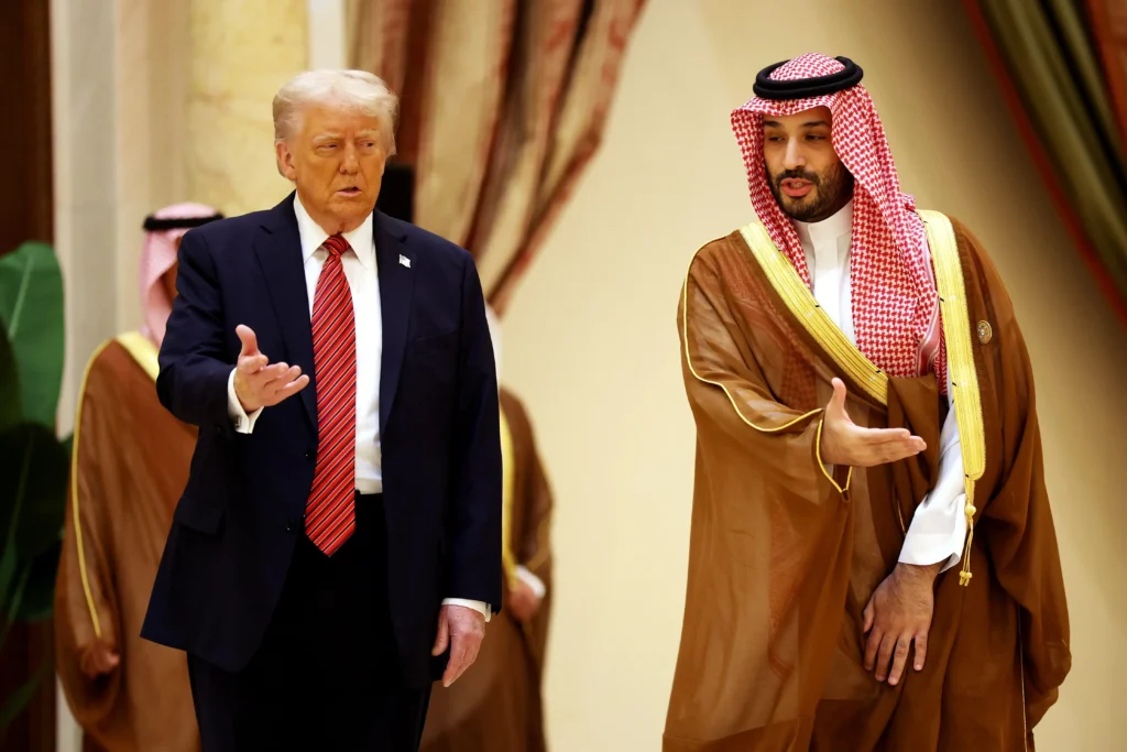 U.S. President Donald Trump in a dark suit and red tie walking alongside Saudi Crown Prince Mohammed bin Salman, who wears traditional Saudi attire, during a meeting in a formal hall.